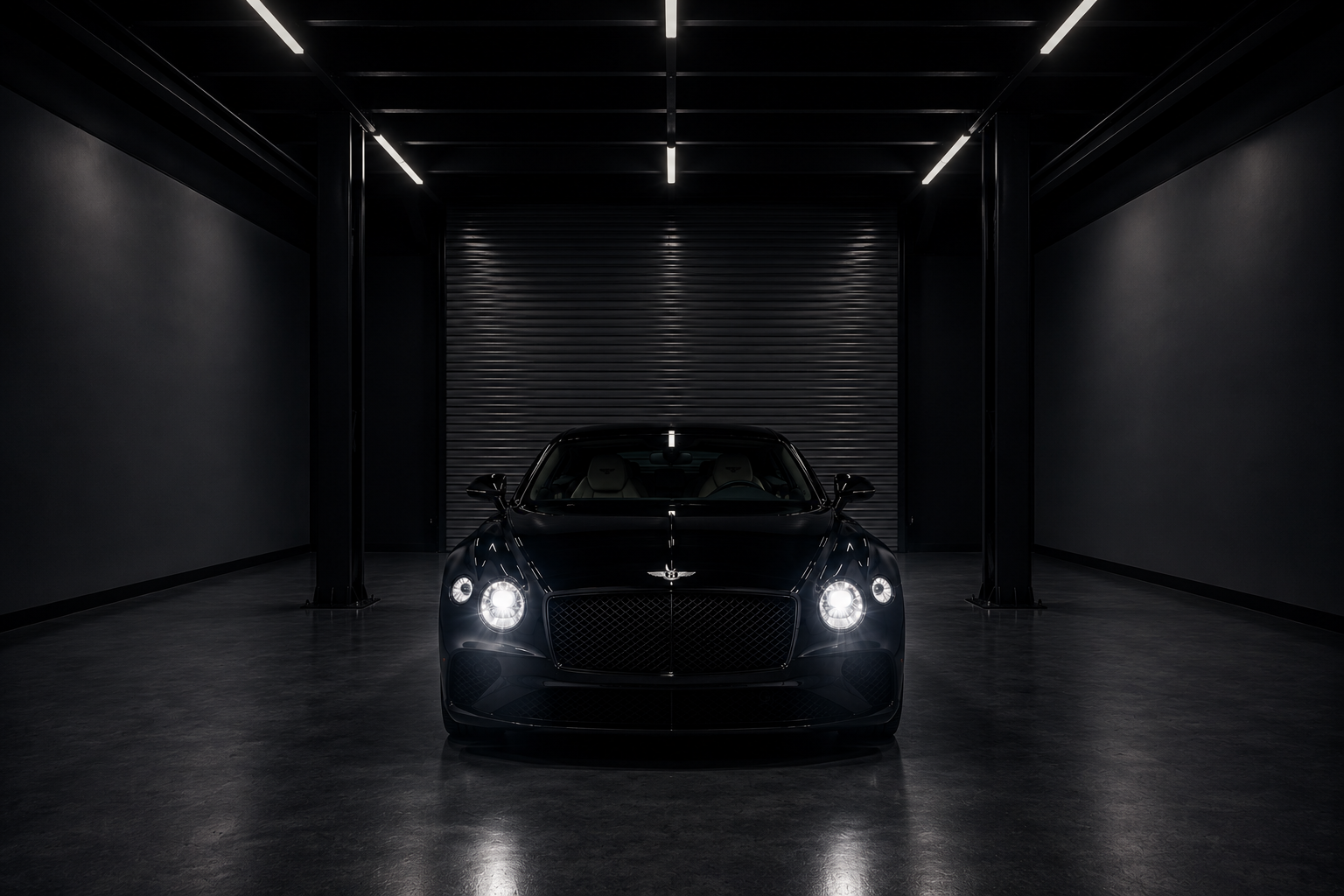 A sleek black luxury coupe sits centered in a dark industrial garage, facing directly toward the camera with bright headlights glowing. The polished concrete floor reflects the overhead strip lighting, while the widened cinematic composition and shadowy surroundings create a dramatic, high-end automotive studio atmosphere.
